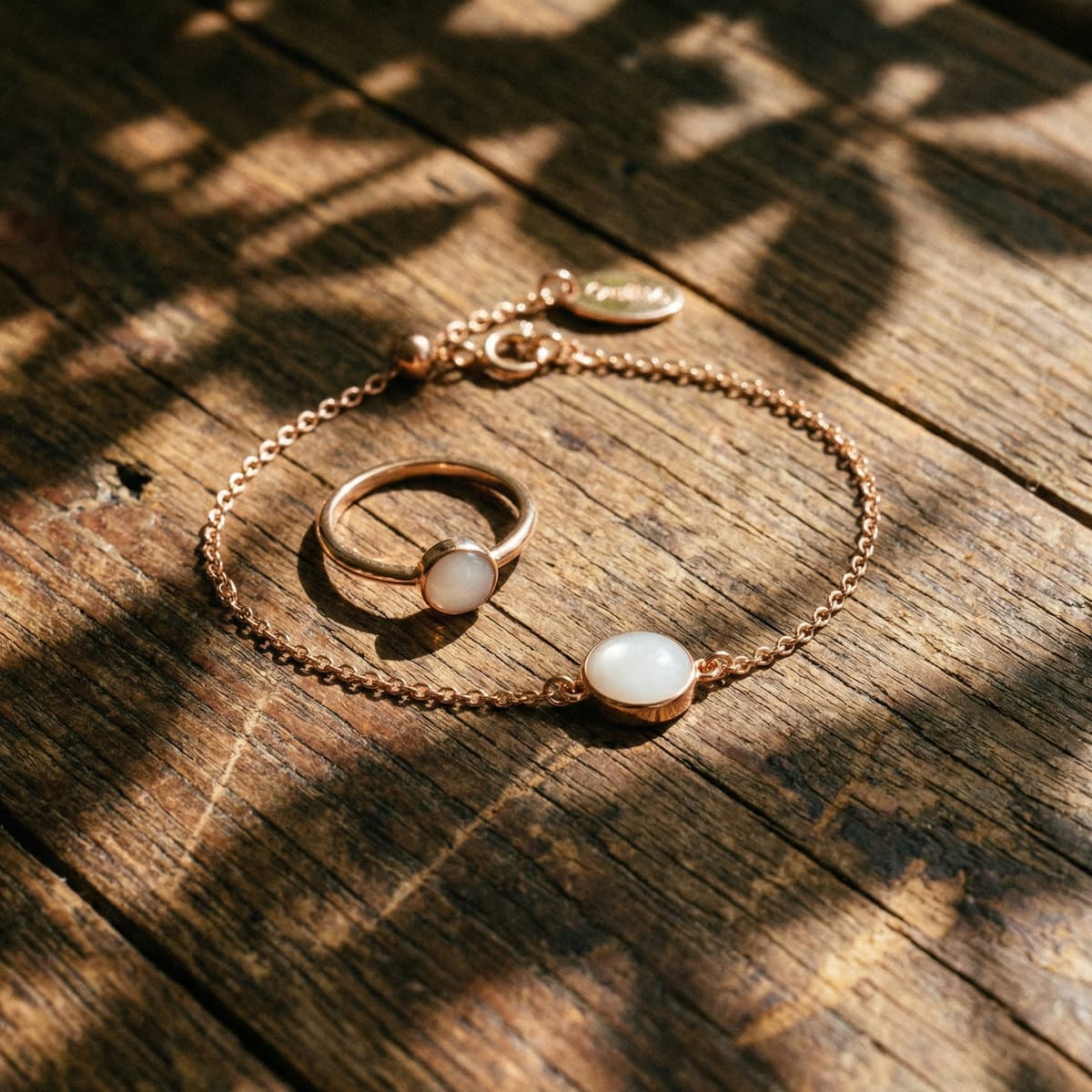 Auf einer rustikalen Holzoberfläche liegen im warmen Sonnenlicht ein zartes, roségoldenes Armband aus Muttermilch mit milchig-weißem Cabochon-Anhänger und ein passender Ring.