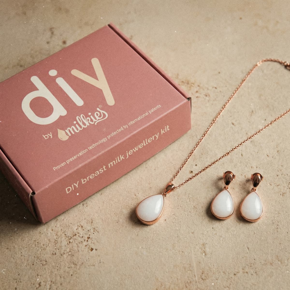 Rosafarbene DIY by MILKIES Brustmilch-Schmuckset-Box als Geschenk für Schwangere neben roségoldener Kette und passenden Tropfen-Ohrringen mit weißem Anhänger.