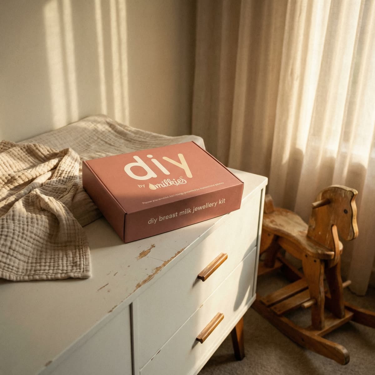 Rosafarbene DIY by MILKIES Brustmilch-Schmuckset-Box als liebevolles Geschenk für Schwangere auf einer weißen Kommode im warmen Sonnenlicht neben einem Schaukelpferd im Kinderzimmer.