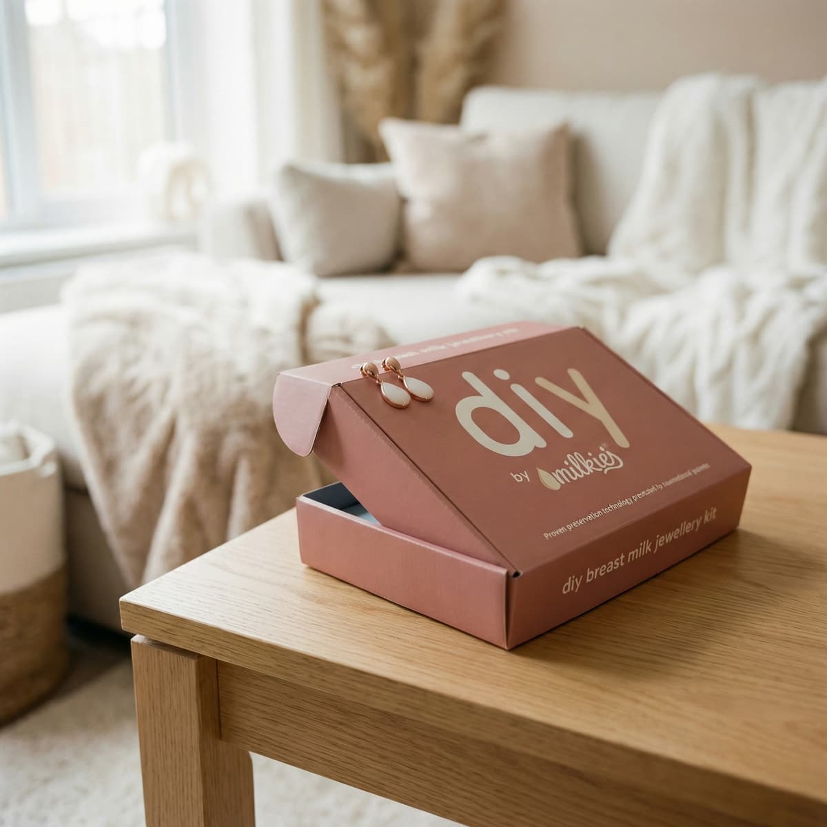 DIY by MILKIES Brustmilch-Schmuckkit-Box auf Holztisch im gemütlichen Wohnzimmer mit roségoldenen Ohrhängern, ideales persönliches Geschenk zum Abstillen für Mütter