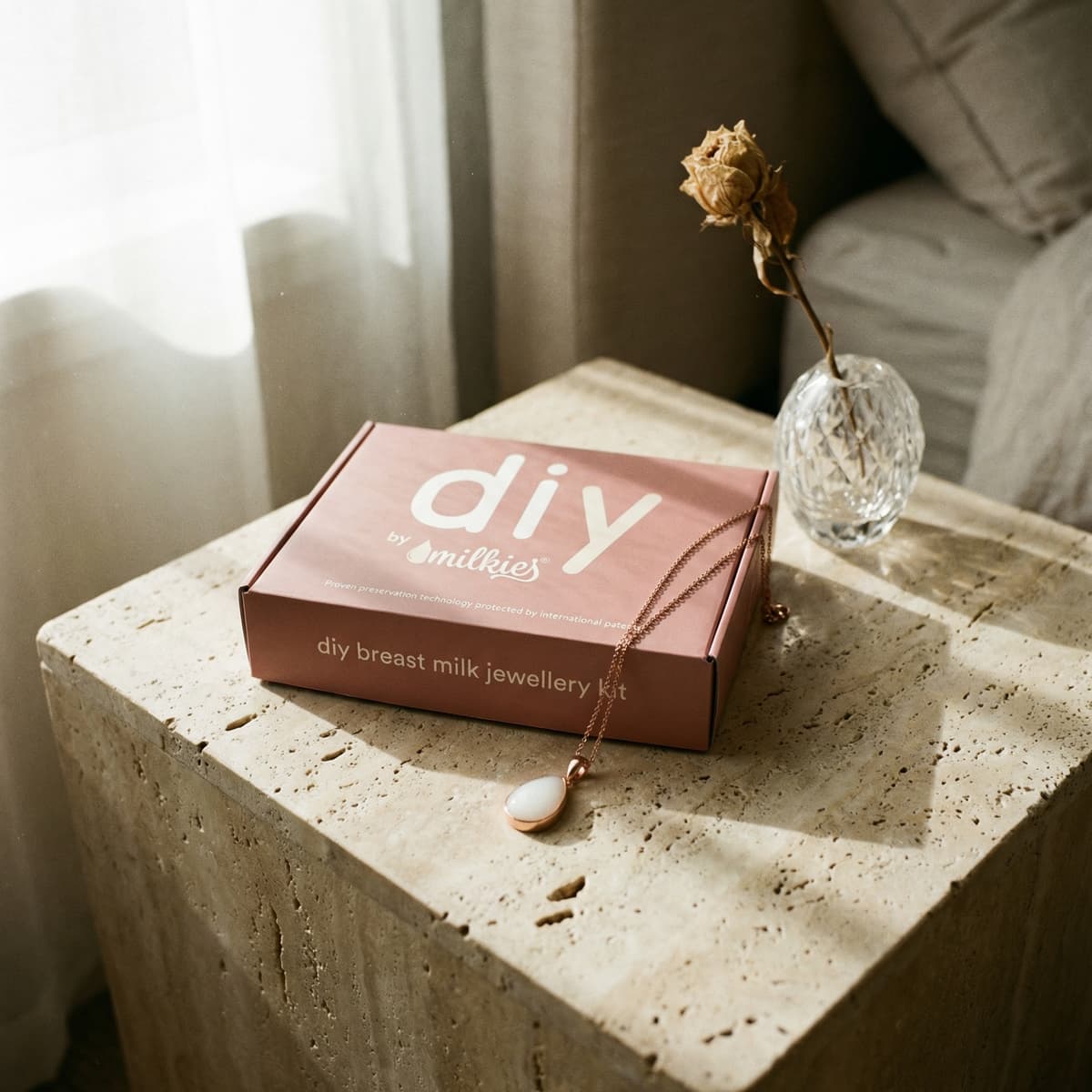 Rosafarbene Box mit DIY by MILKIES Brustmilch-Schmuckset als persönliches Geschenk zur Geburt, Kette auf Steinblock neben Glasvase im warmen Sonnenlicht.