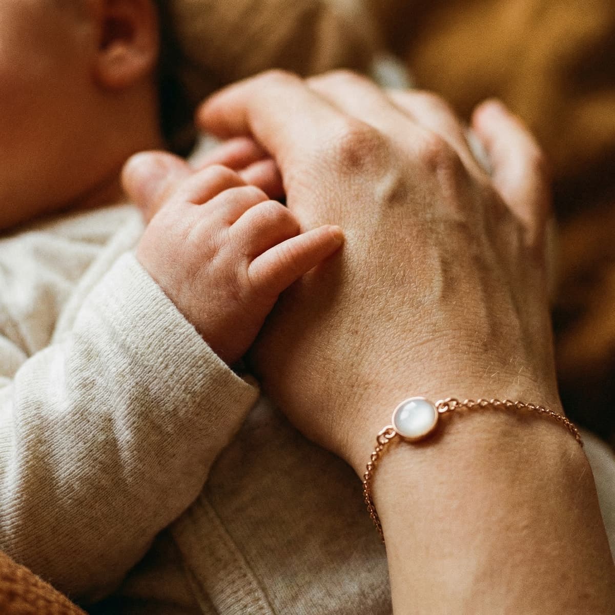 Nahaufnahme einer Mutterhand, die die Hand ihres Babys hält, mit zartem roségoldenem Armband und milchig-weißem Stein als Muttermilch Armband.