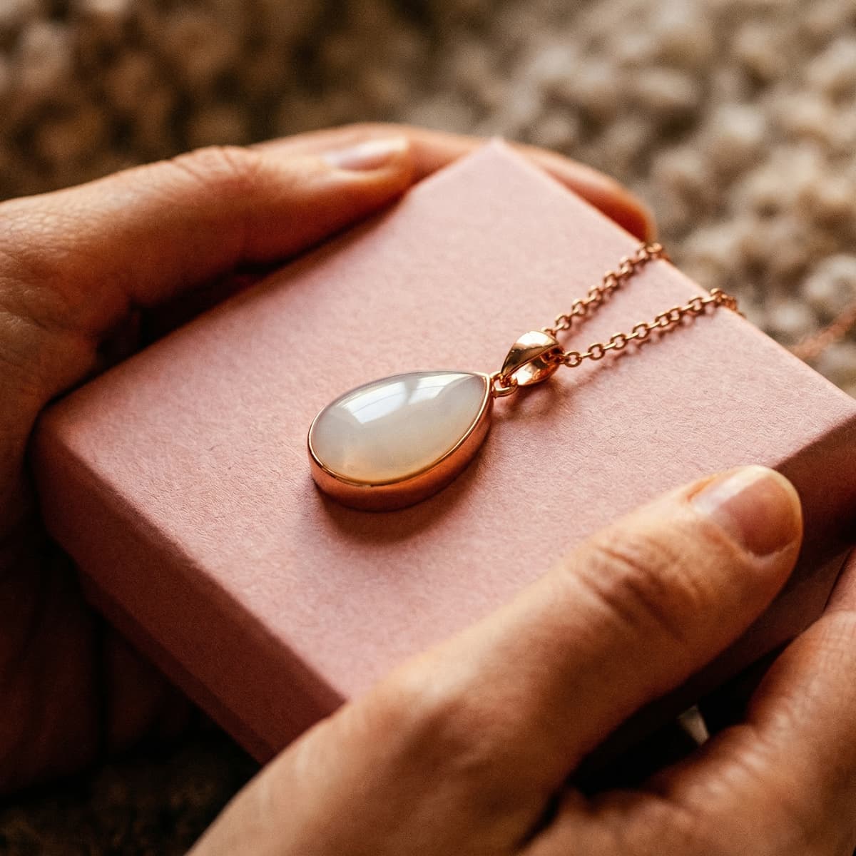 Elegante Muttermilch Halskette in Roségold mit tropfenförmigem milchig-weißem Anhänger, in Händen über einer rosa Schmuckbox – emotionaler Grund, das DIY by MILKIES Kit zu wählen und den Erinnerungsstein zuhause selbst zu gestalten.