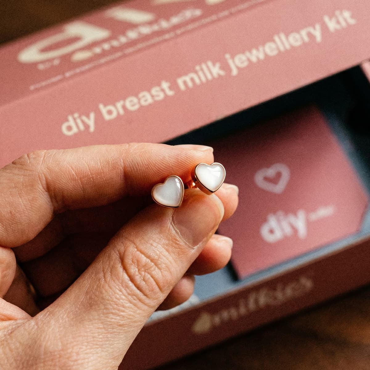 Eine Hand hält zarte, herzförmige Muttermilch Ohrringe in Goldoptik vor der Verpackung eines DIY-by-MILKIES Breastmilk-Jewellery-Kits.