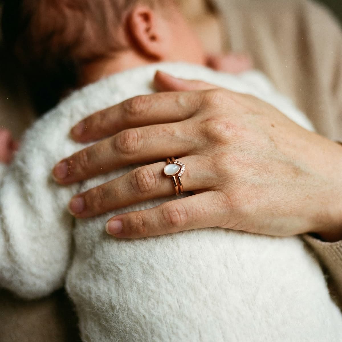 Hand einer Mutter, die ein Baby hält, mit elegantem Muttermilch Ring in Roségold als persönliches Erinnerungsstück – zeigt, warum das DIY by MILKIES Kit eine besondere Idee für ein DIY-Andenken ist.