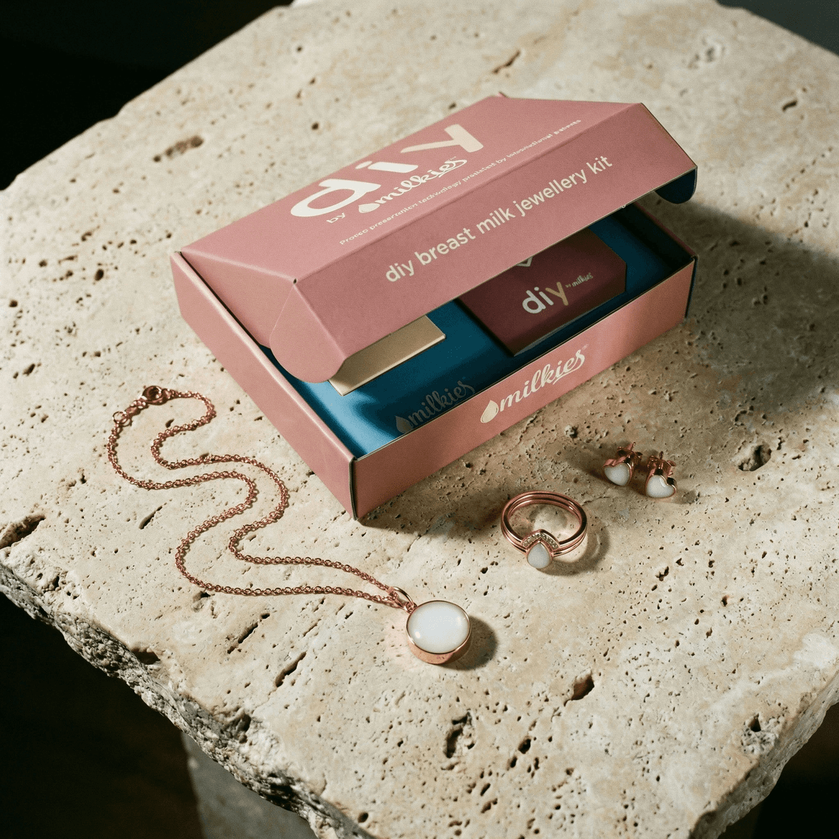 Rosafarbene Box mit offenem Muttermilch Schmuck DIY Set von DIY by MILKIES auf Steinoberfläche, daneben roségoldene Halskette, Ring und Ohrstecker mit milchig-weißem Schmuckstein.