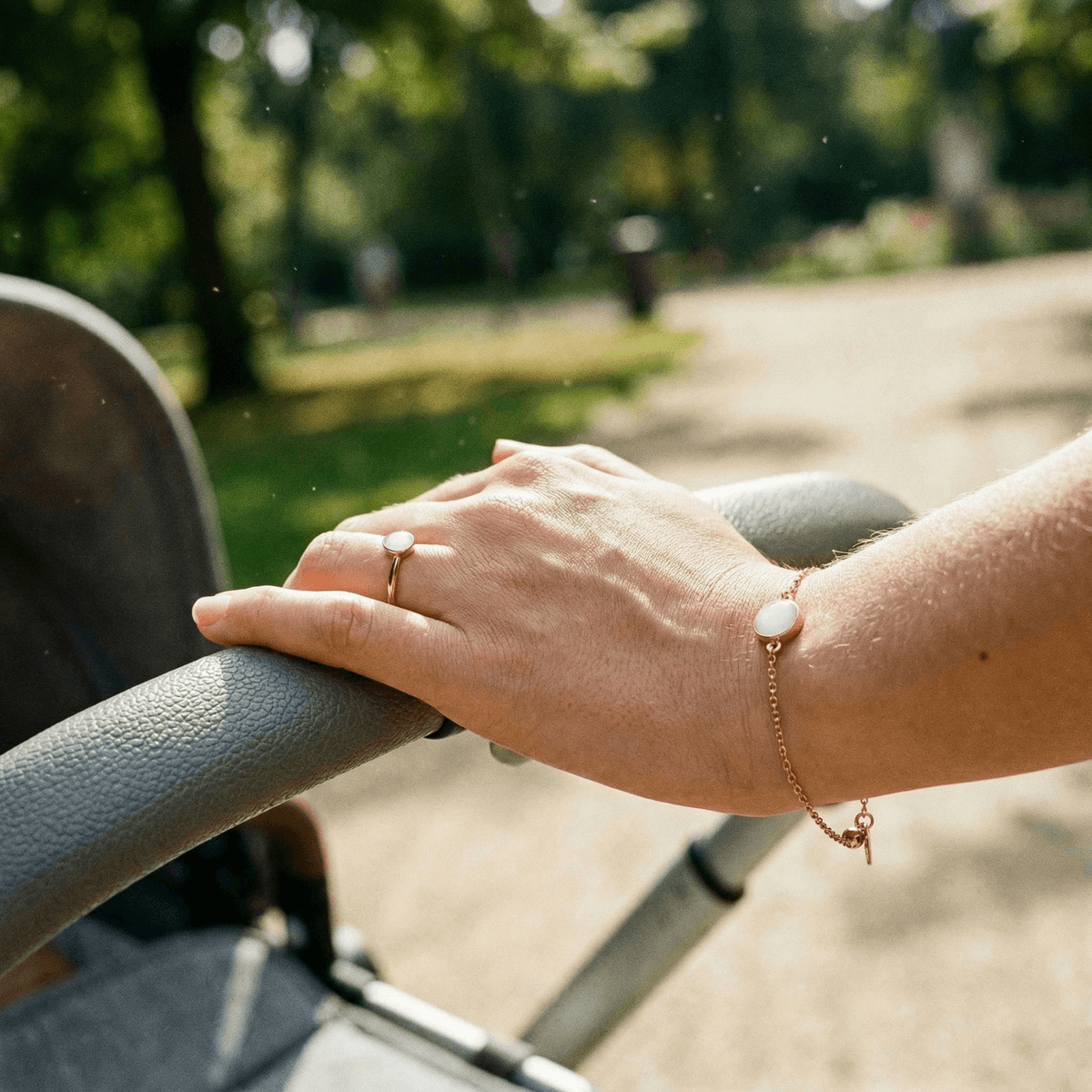 Hand einer Mutter am Kinderwagen im Park mit zartem Muttermilchschmuck: goldener Ring und Armband mit milchig-weißem Stein als Erinnerungsschmuck.