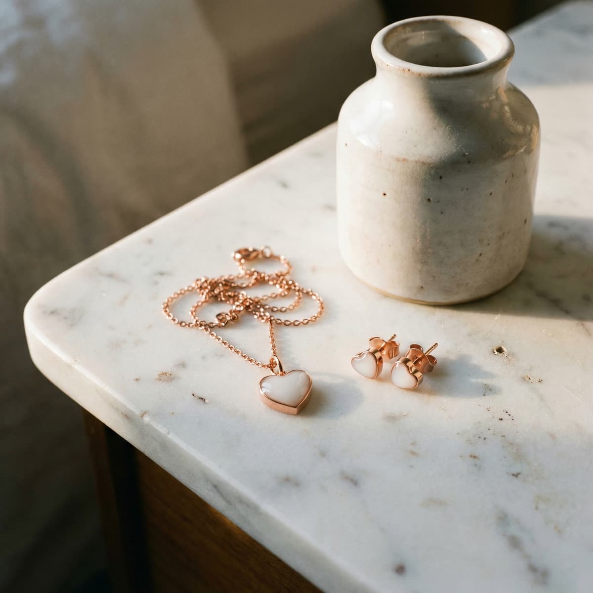 Roségoldenes Schmuck-Set mit Herz-Anhänger-Collier und passenden Herz-Ohrsteckern als Muttermilchschmuck DIY Set, dekorativ auf heller Marmortischplatte neben weißer Keramikvase im warmen Tageslicht arrangiert.