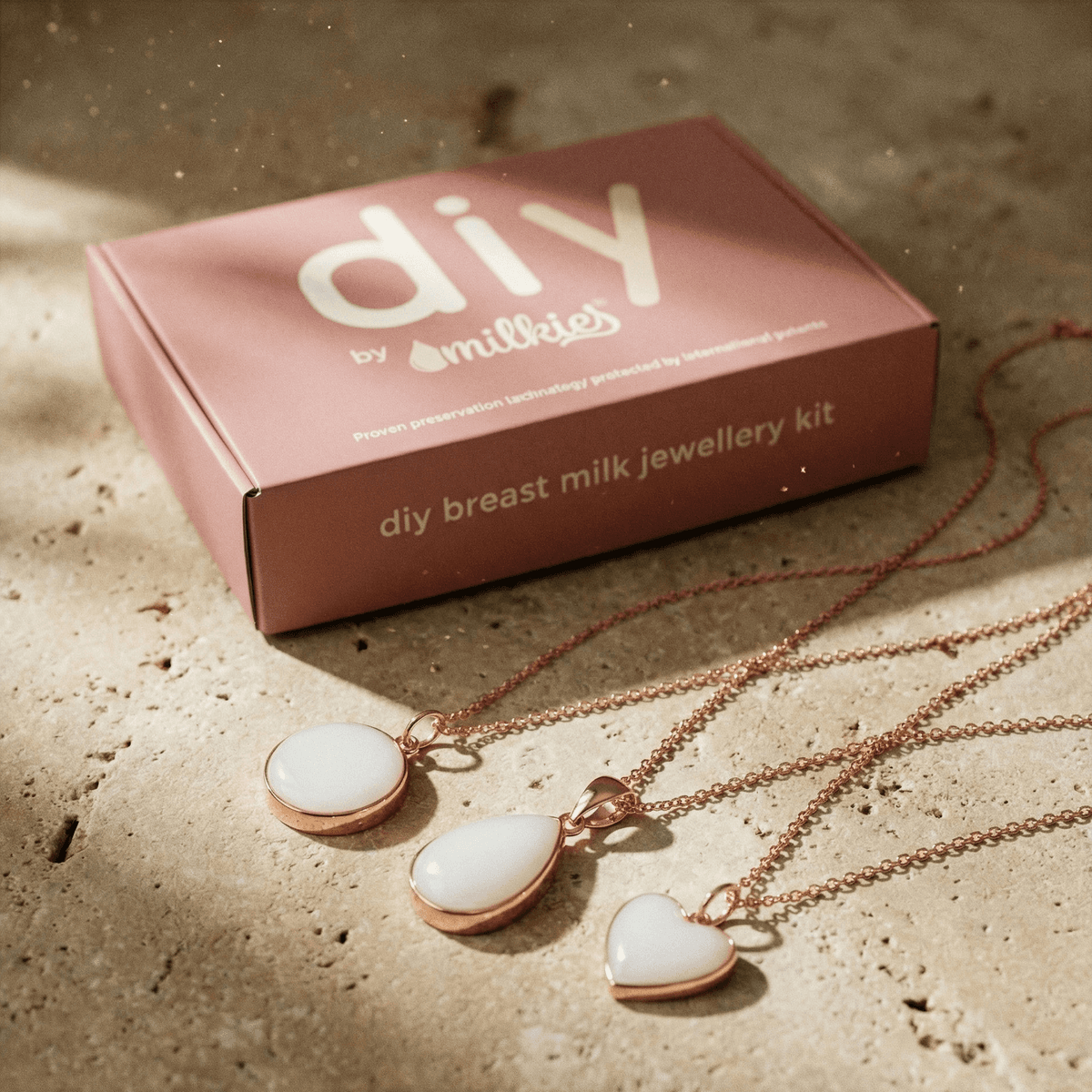 Rosafarbene Schmuckkit-Box „diy breast milk jewellery kit“ von DIY by MILKIES mit drei roségoldfarbenen Anhängern in Weiß (rund, tropfenförmig, Herz) – elegantes Set DIY Muttermilch Kette auf Steinoberfläche.