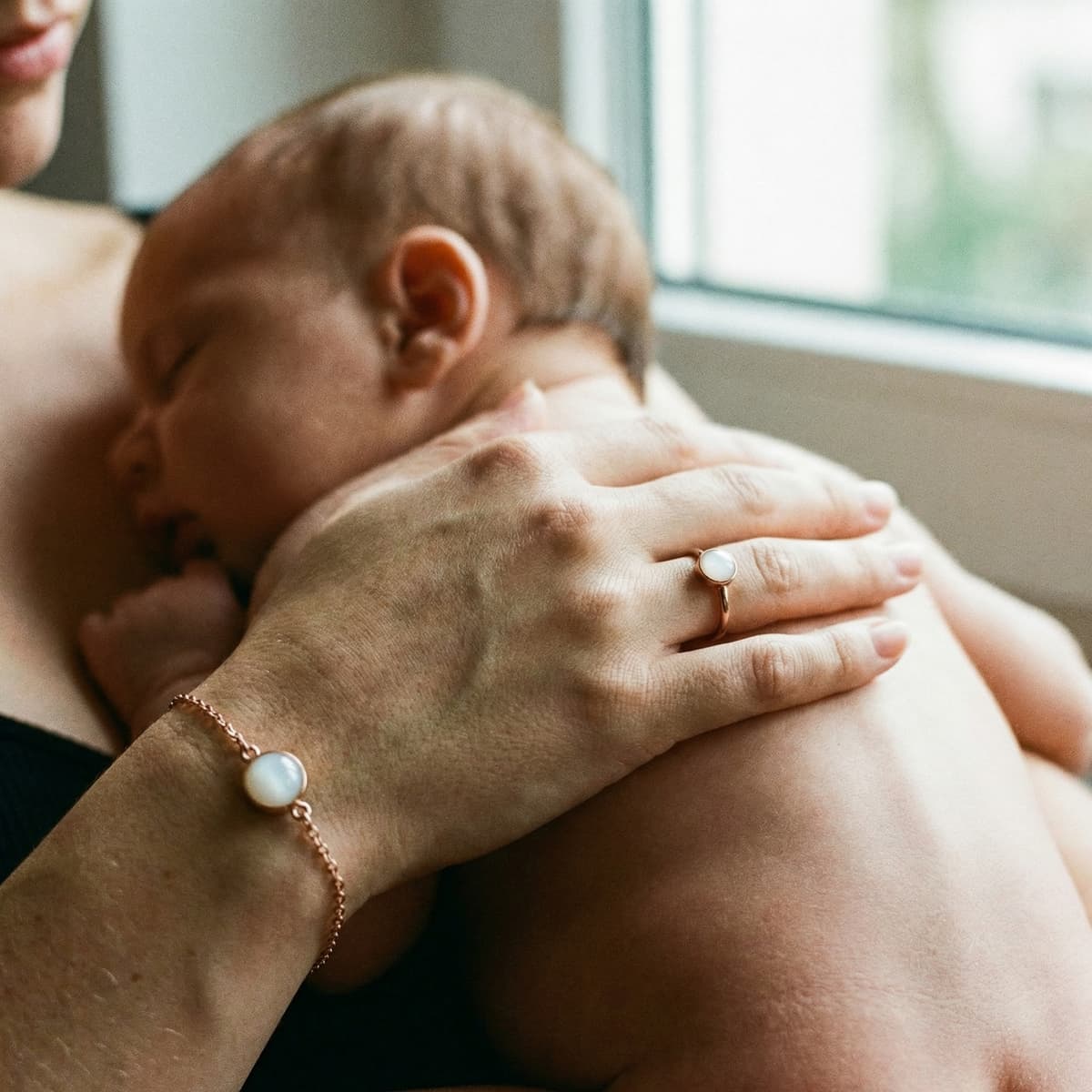 Mutter hält ihr Baby am Fenster und trägt ein roségoldenes Armband mit milchig-weißem Stein – Inspiration, um ein Armband mit Muttermilch selber machen zu können und warum ein DIY-Kit ein persönliches Andenken zu Hause ermöglicht.
