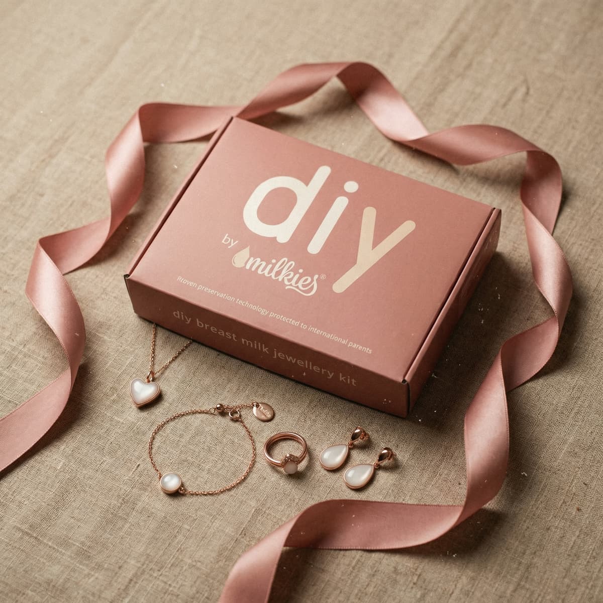 Stilvolle Geschenkidee als Geschenk für frischgebackene Mama: DIY by MILKIES Brustmilch-Schmuckset mit rosafarbenem Kit, Satinband und roségoldfarbenem Schmuck (Kette, Armband, Ring, Ohrringe) auf Stoffhintergrund.