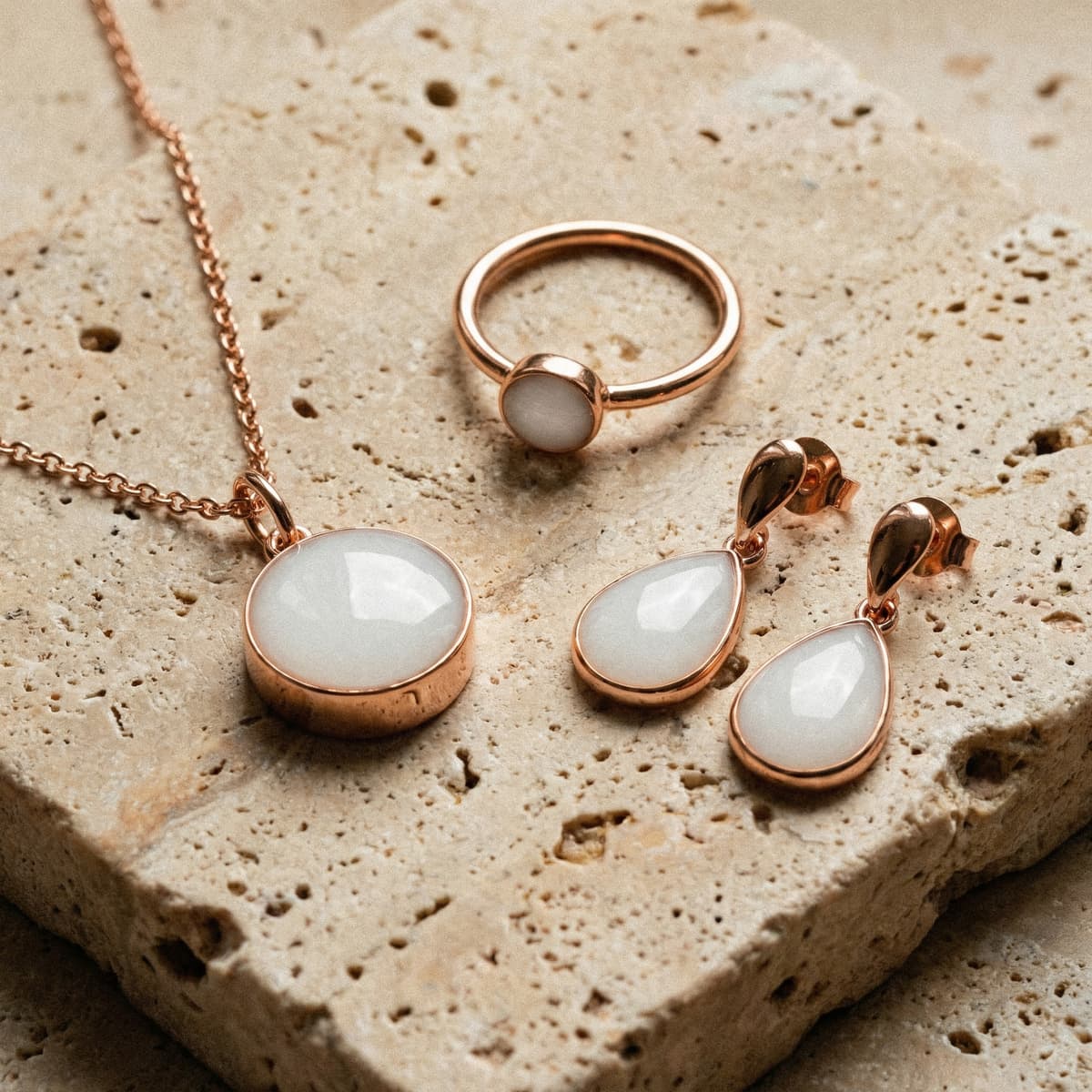 Roségoldenes Schmuck-Set mit Anhänger, Ring und Tropfen-Ohrringen auf einer Steinplatte, inspiriert von einem Resin Set für Muttermilchschmuck mit milchig-weißen Resin-Steinen.